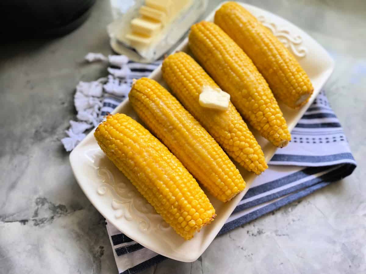 5 corn on the cobs on a long white dish with a pad of butter on top with slices of butter on the side