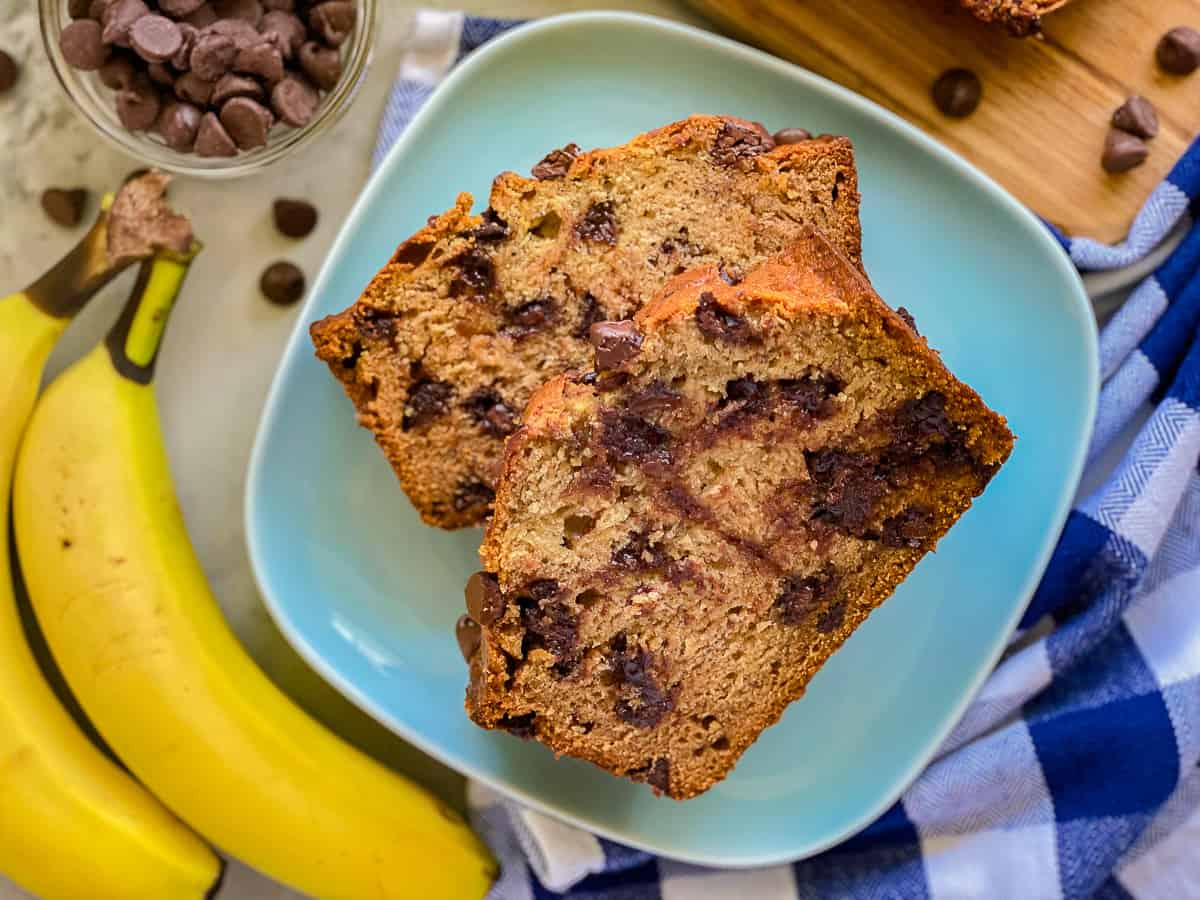 Blue plate with slices of banana bread with chocolate chips with yellow bananas and chocolate chips next to the plate.