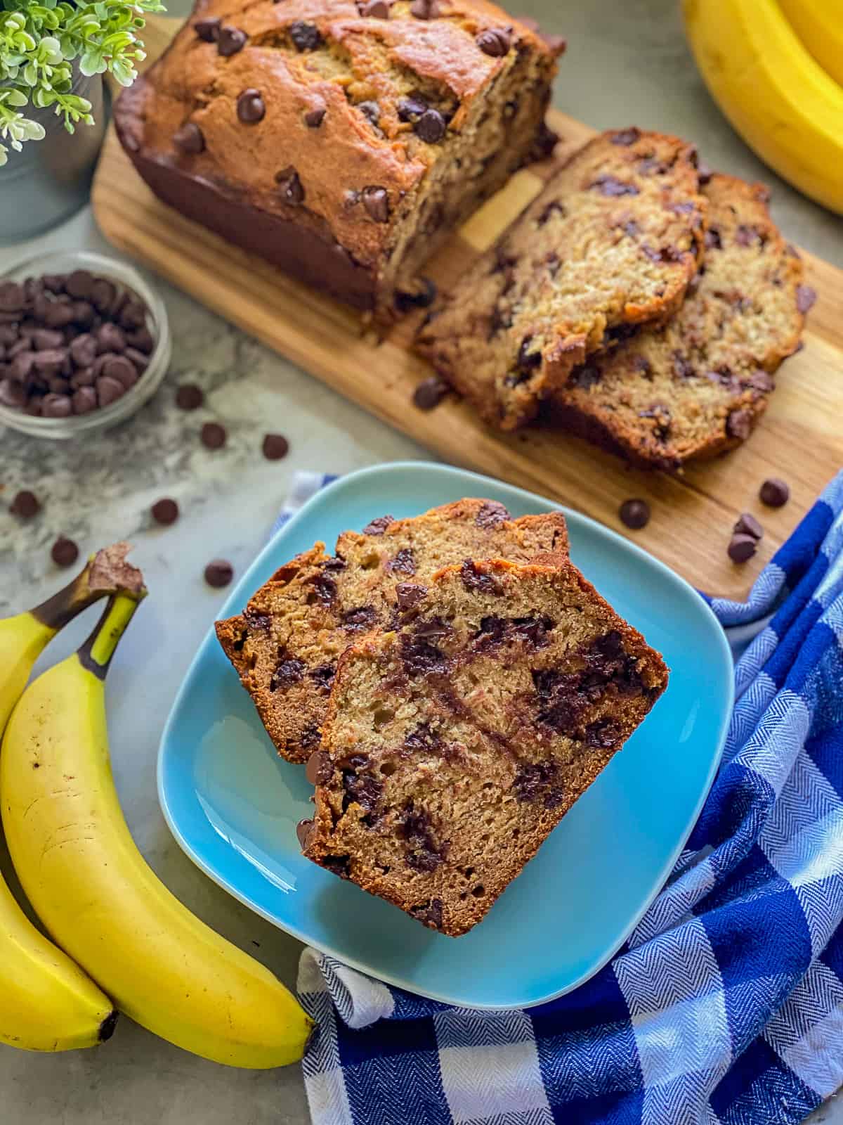 Blue plate filled with two slices of chocolate chip banana bread with yellow bananas next to the plate.