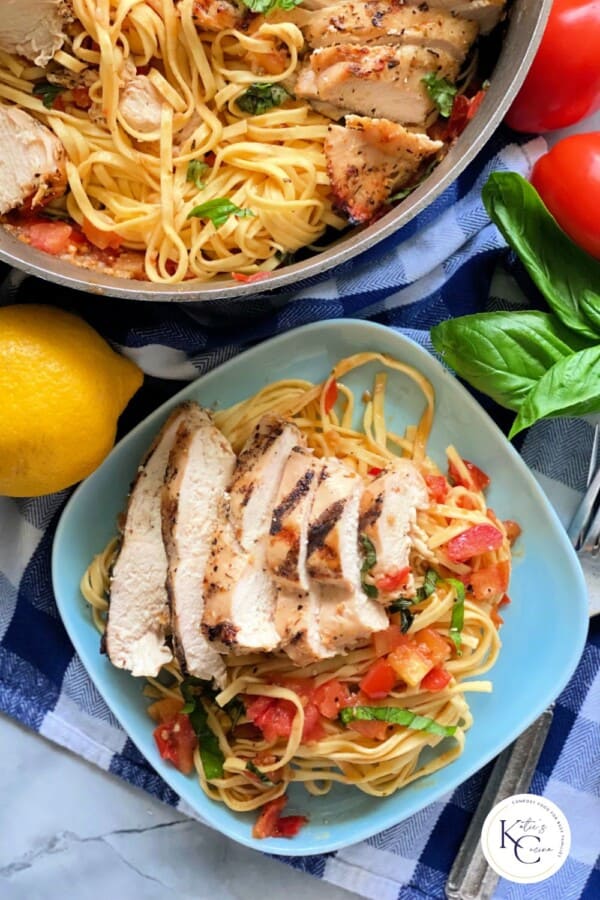 Blue plate filled with pasta and sliced chicken sitting on a blue and white checkered cloth with a pot filled with pasta and chicken with logo on bottom right corner.