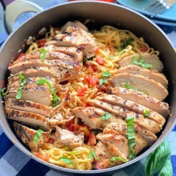 Skillet filled with pasta, tomatoes, basil, and sliced chicken on a blue and white checkered cloth.