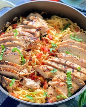 Skillet filled with pasta, tomatoes, basil, and sliced chicken on a blue and white checkered cloth.