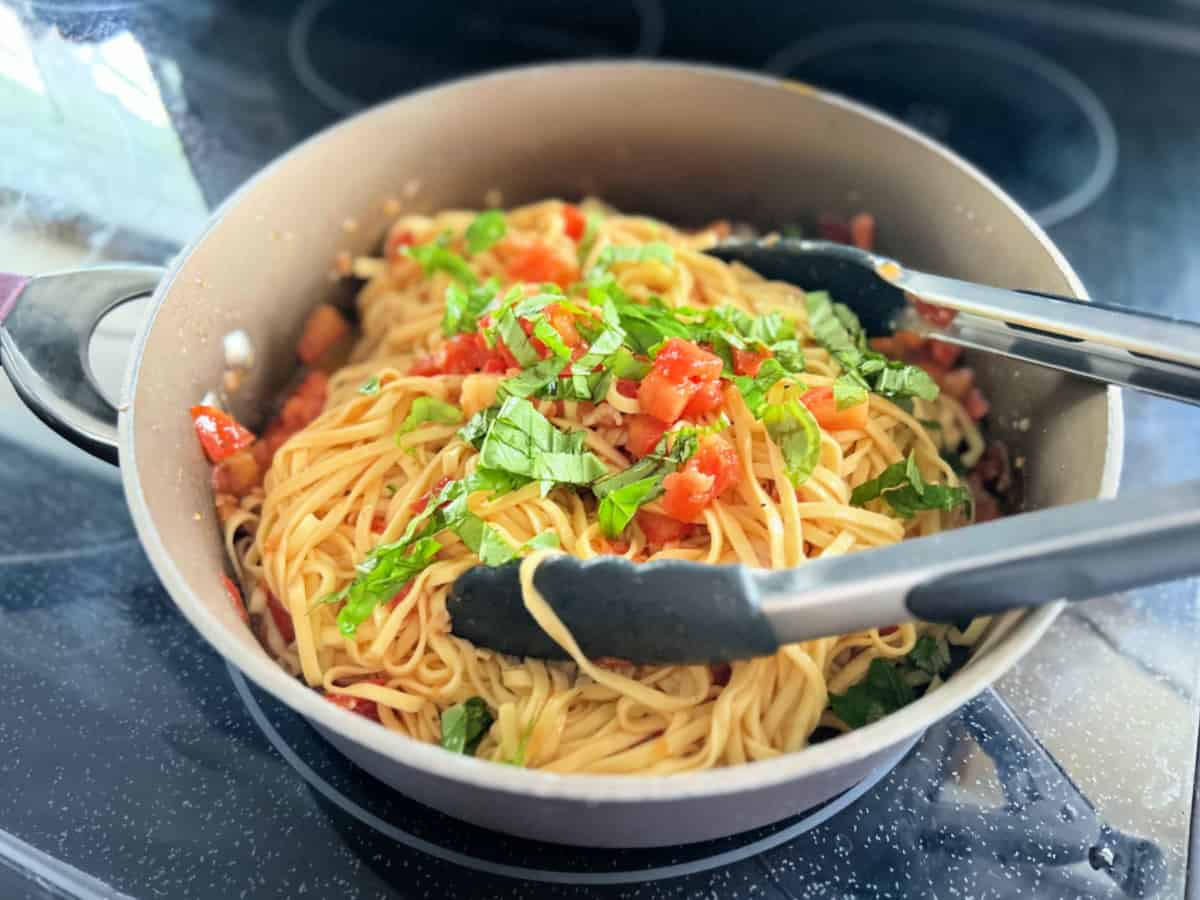 Black stove top with a brown pot with pasta, tomatoes, fresh basil, and tongs.