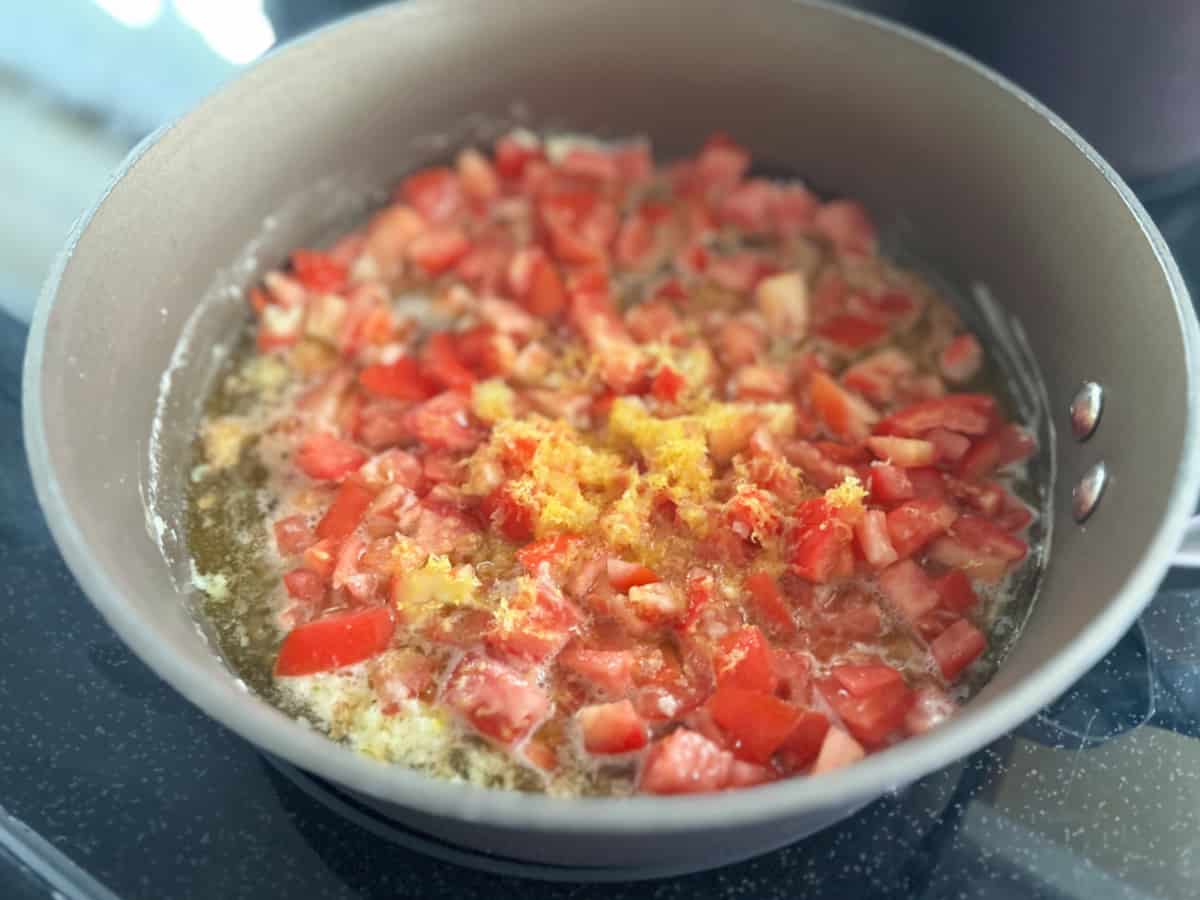 Brown pot with tomatoes, oil, butter, and grated lemon on a stove top.