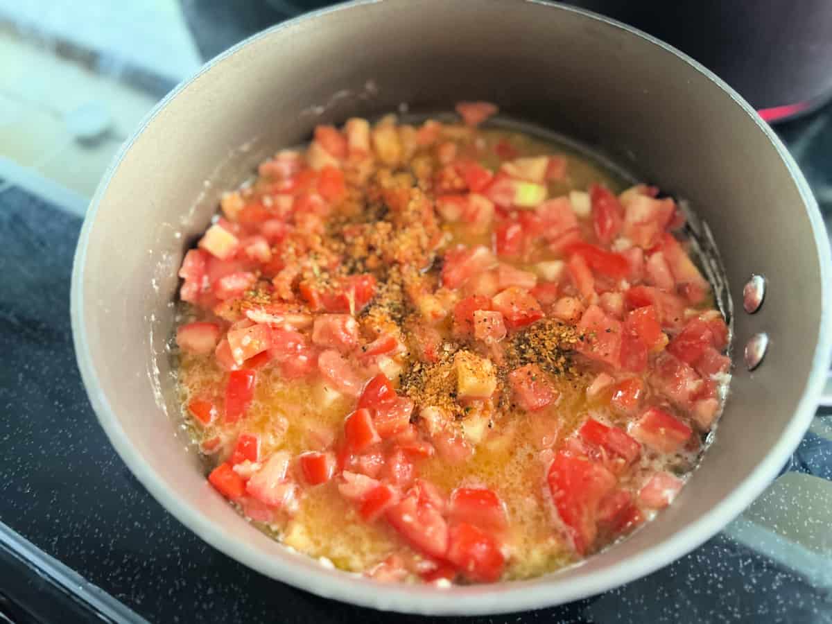 Brown pot filled with diced tomatoes, seasoning, and butter/oil on a black stove top.