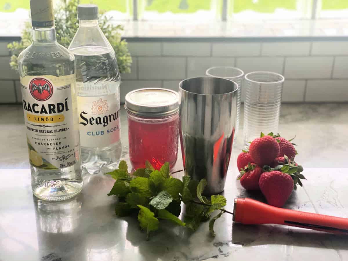 Ingredients on marble countertop: bacardi limon, club soda, strawberry simple syrup, strawberries, and mint. 