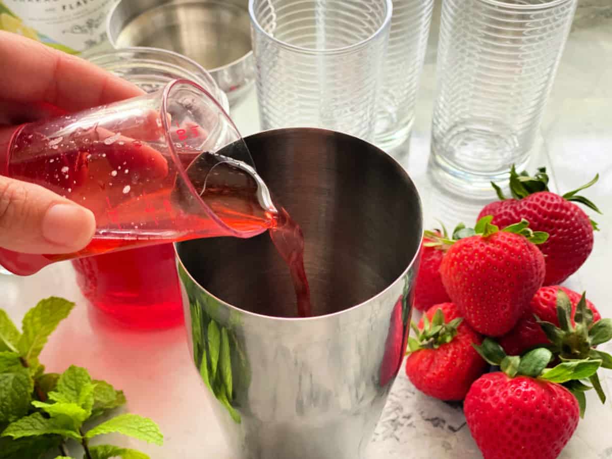 Metal shaker with female hand pouring strawberry simple syrup.