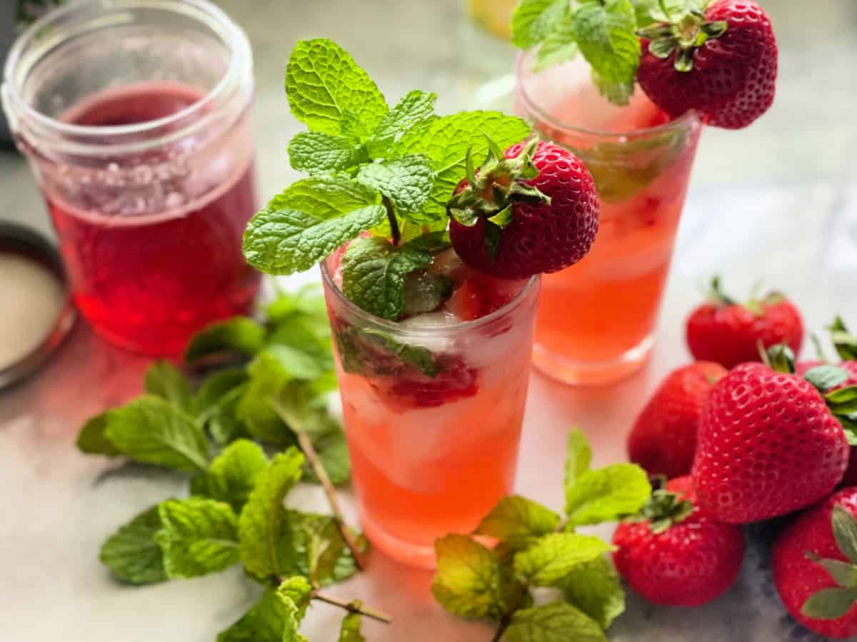Two glasses filled with a pink drink and mint and strawberry as garnish with a mason jar filled with a pink liquid.