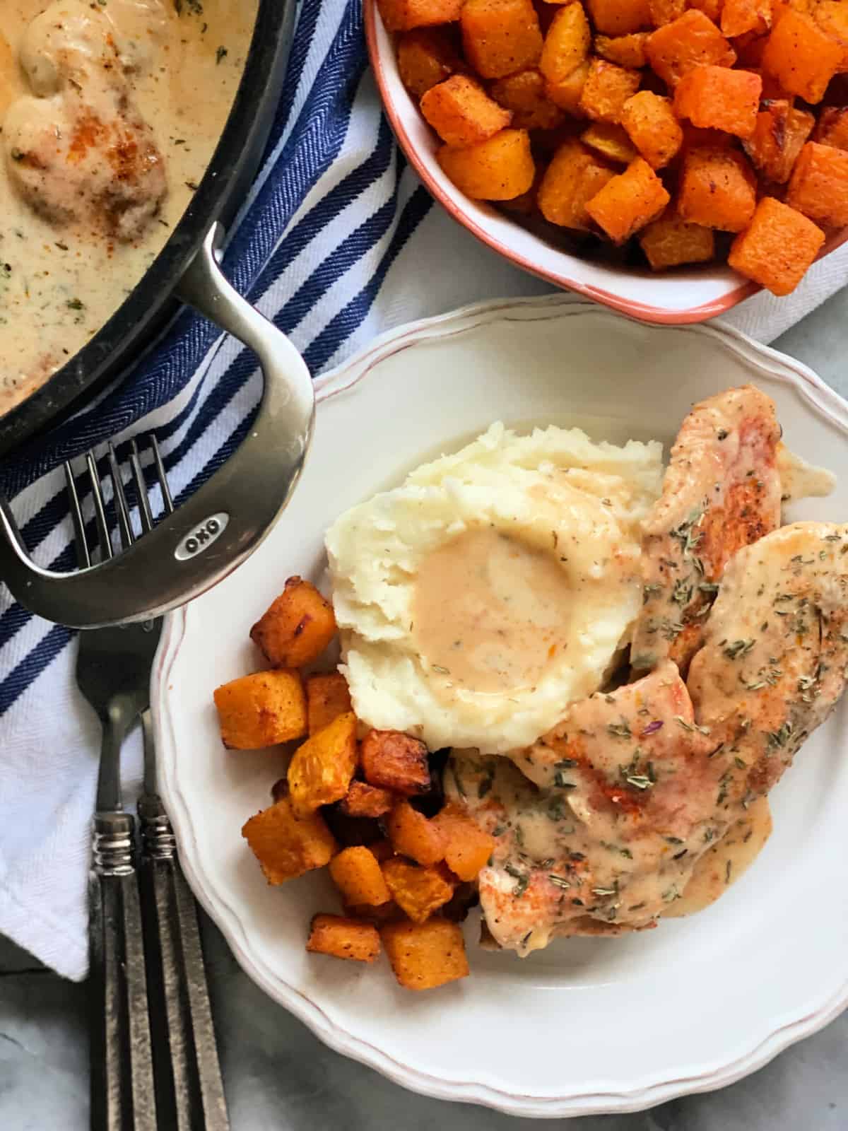Plate with mashed potatoes, turkey cutlets, and butternut squash and air fryer butternut squash in a dish on a blue and white striped cloth and two forks.