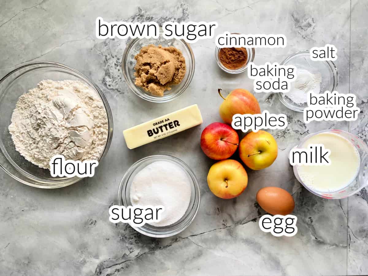 ingredients on marble counter: flour, sugar, brown sugar, sugar butter, apples, cinnamon, egg, baking soda, baking powder, salt, milk.