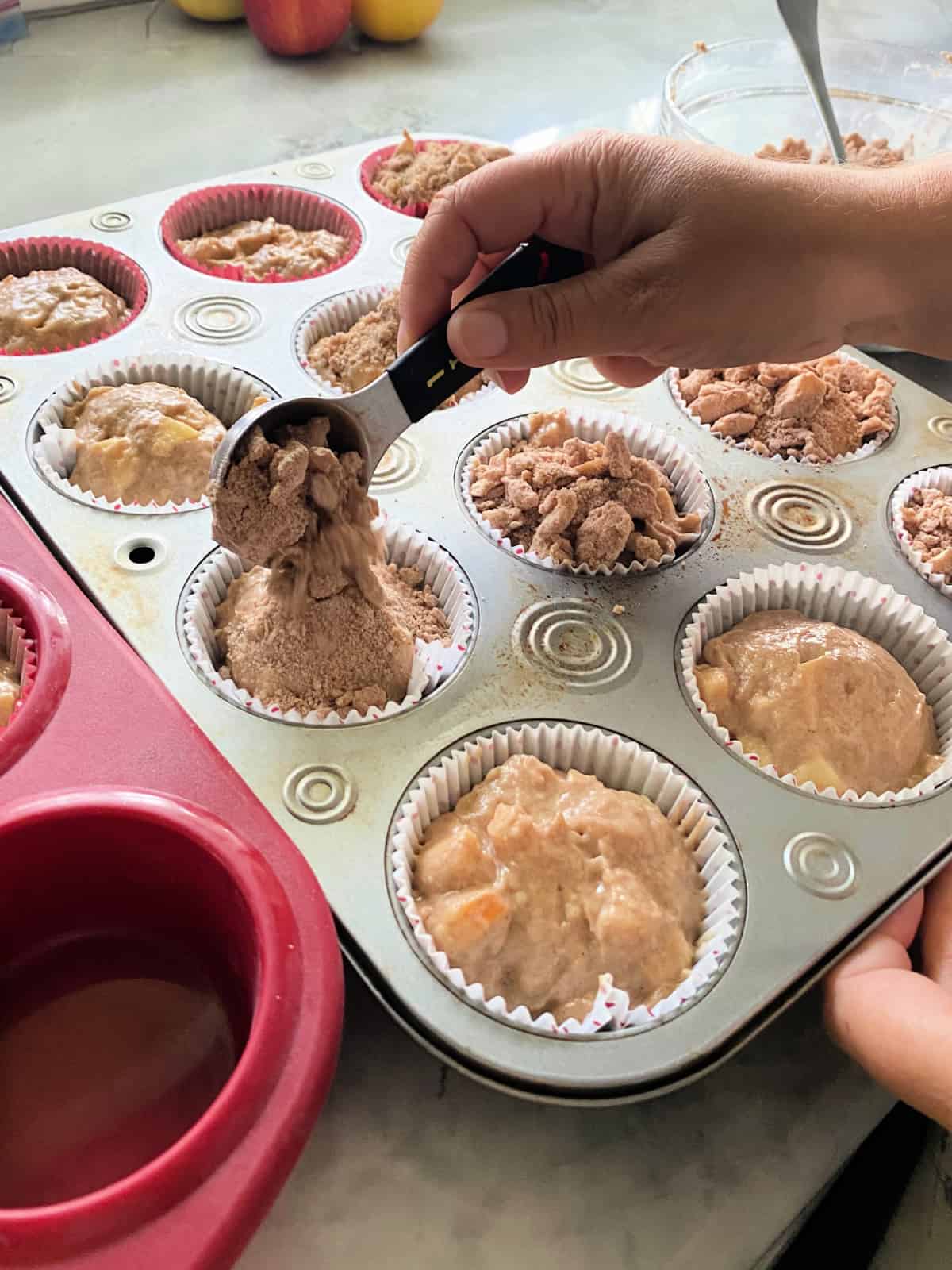 silver cupcake tin with raw muffin batter and the cumb topping being poured.