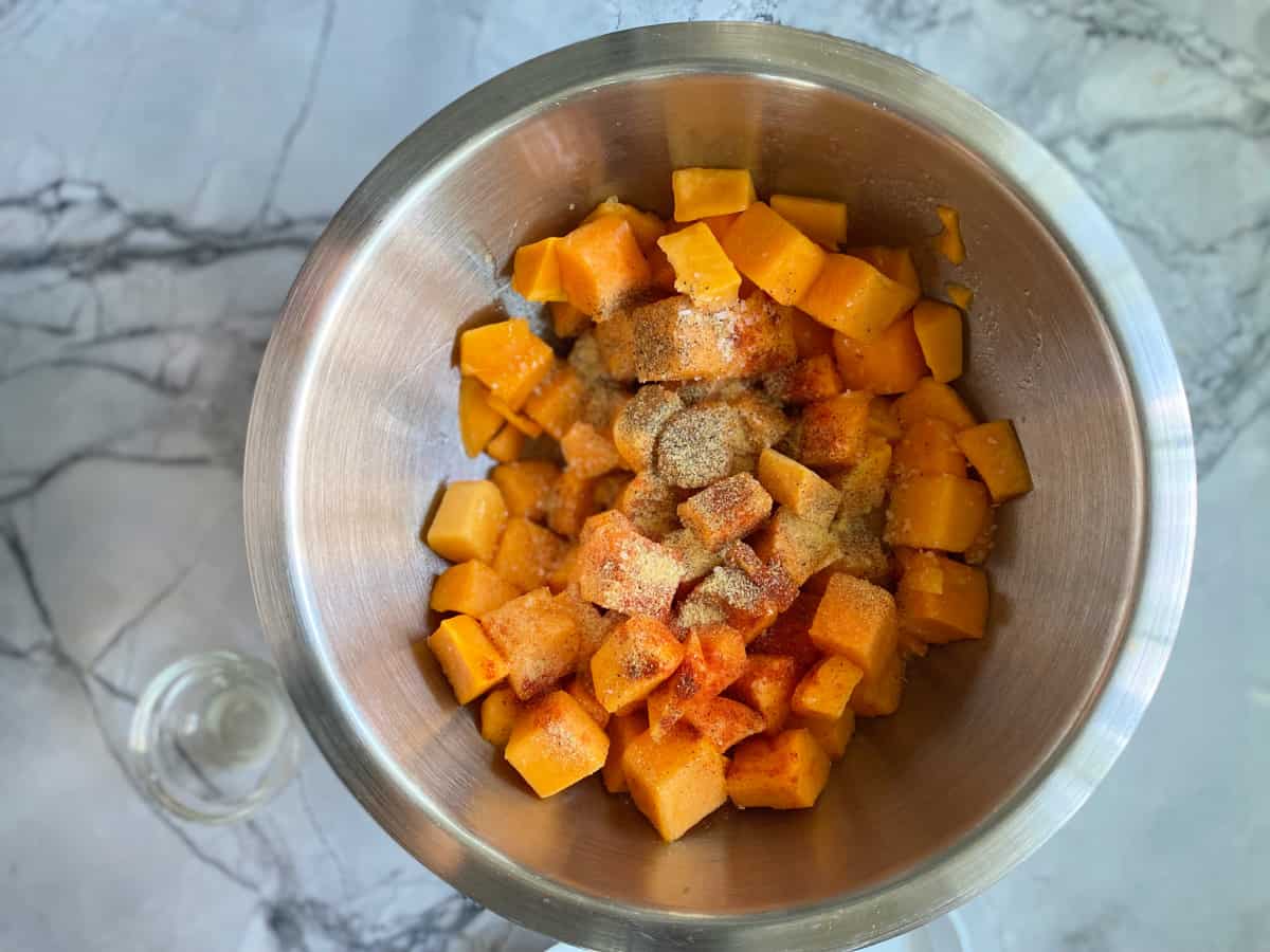 raw diced butternut squash and seasoning in a stainless steel bowl on a marble counter.