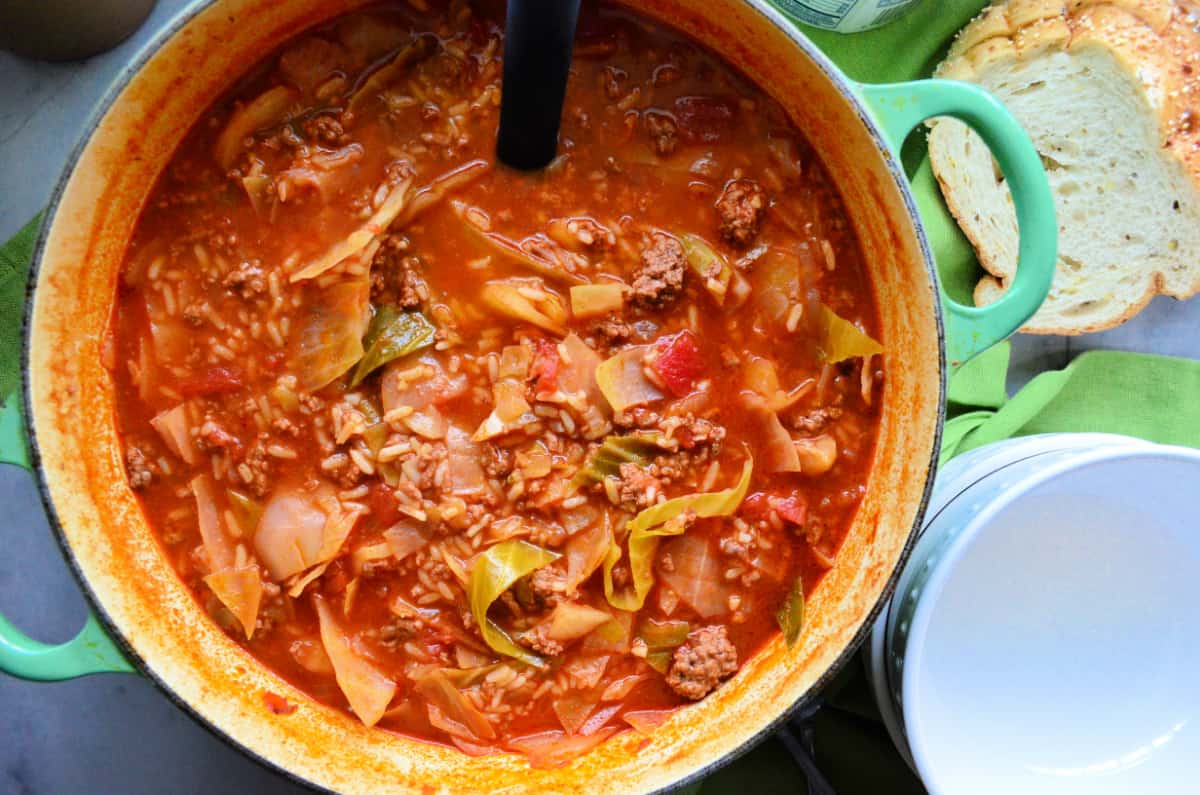 a pot full of cabbage soup on green fabric and a ladle and slices of bread and white bowls in the background.