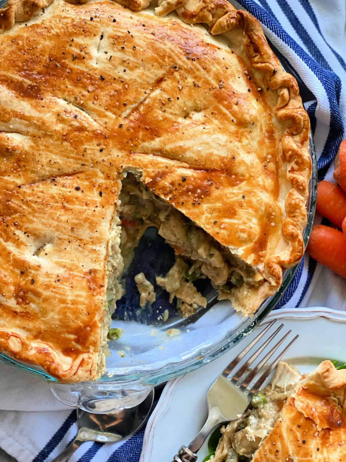 chicken pot pie with one slice cut out on a blue and white stripped cloth, a plate with a slice of chicken pot pie, a spoon, a fork, and carrots