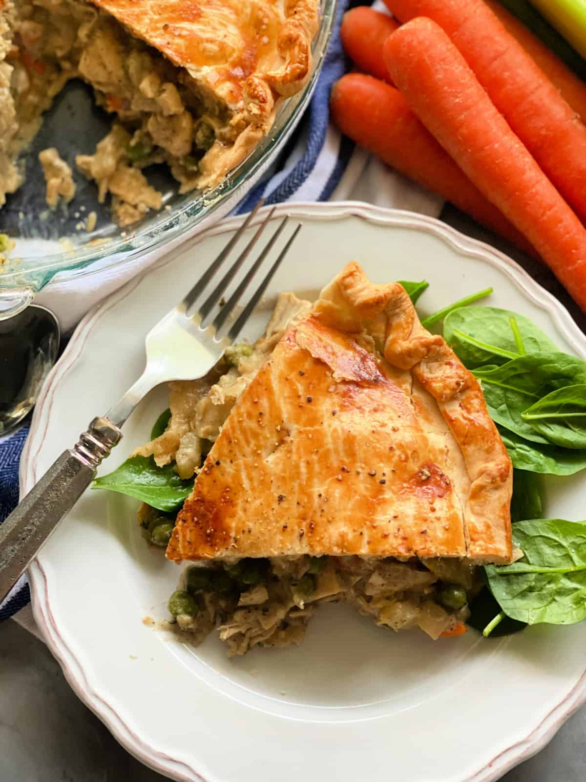 white plate with a slice of chicken pot pie on a blue and white stripped cloth, a spoon, a fork, and carrots in the background.