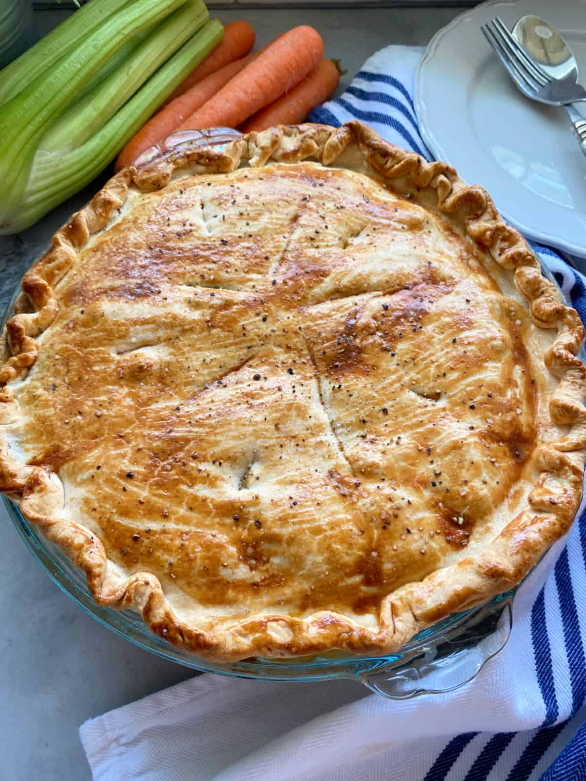 chicken pot pie on a blue and white striped cloth with celery, carrots, plate, fork, and spoon in the background