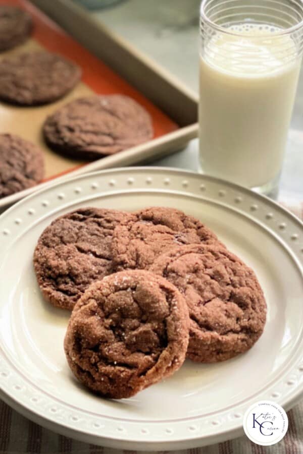 4 chocolate sugar cookies on a white plate with a pink striped white cloth in the background with a glass of milk and 4 baked cookies on a cookie sheet.