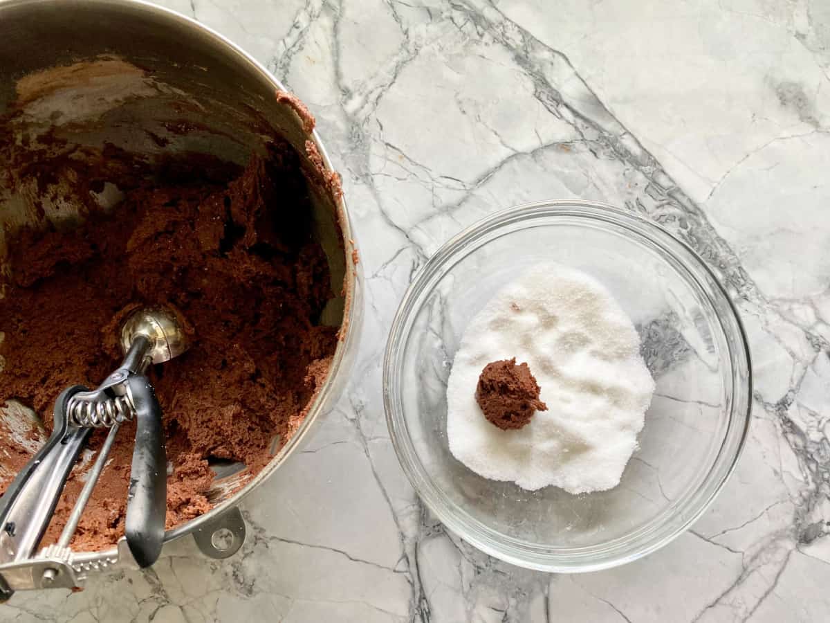 mixing bowl with chocolate sugar cookie dough and a scoop and white sugar and one ball of cookie dough in a clear bowl on a gray marble counter.