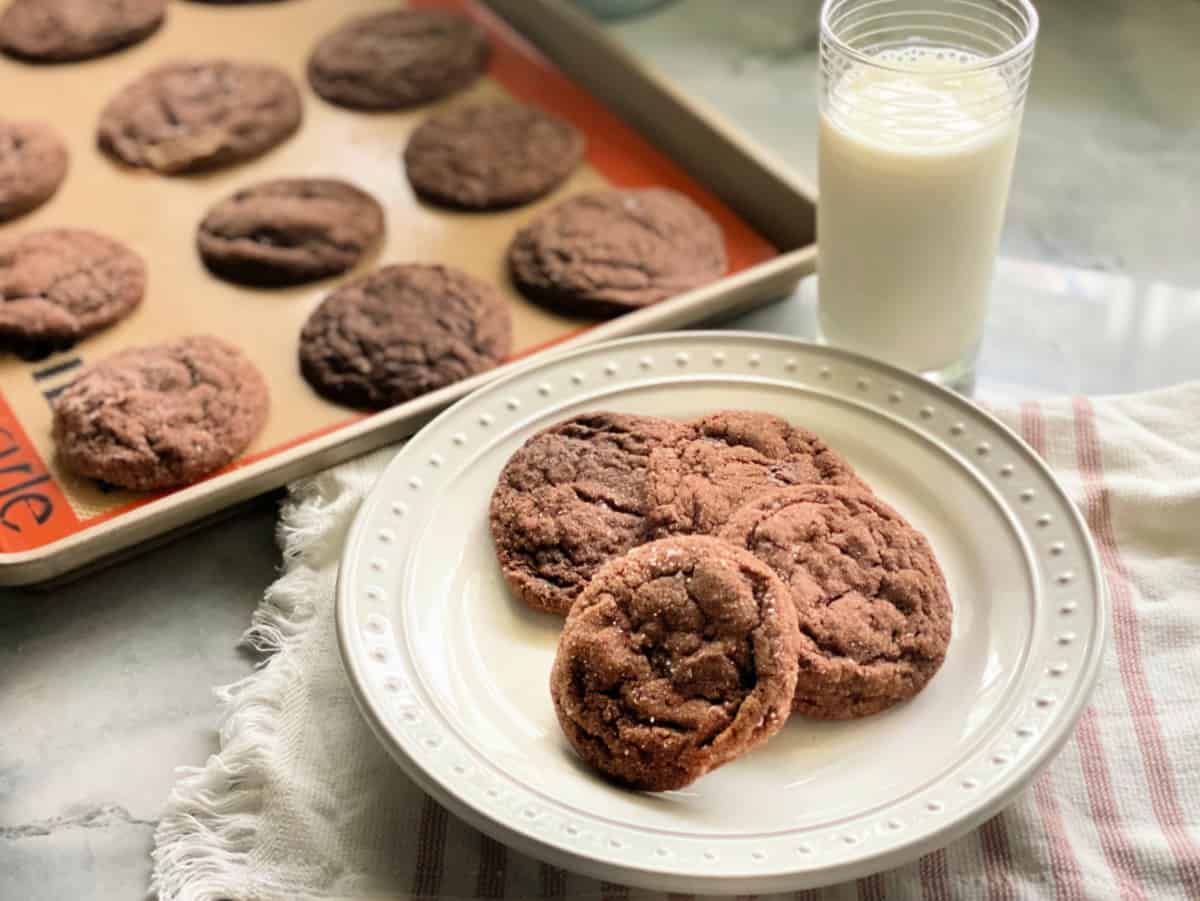 4 chocolate sugar cookies on a white plate with a pink striped white cloth in the background with a glass of milk and 10 baked cookies on a cookie sheet
