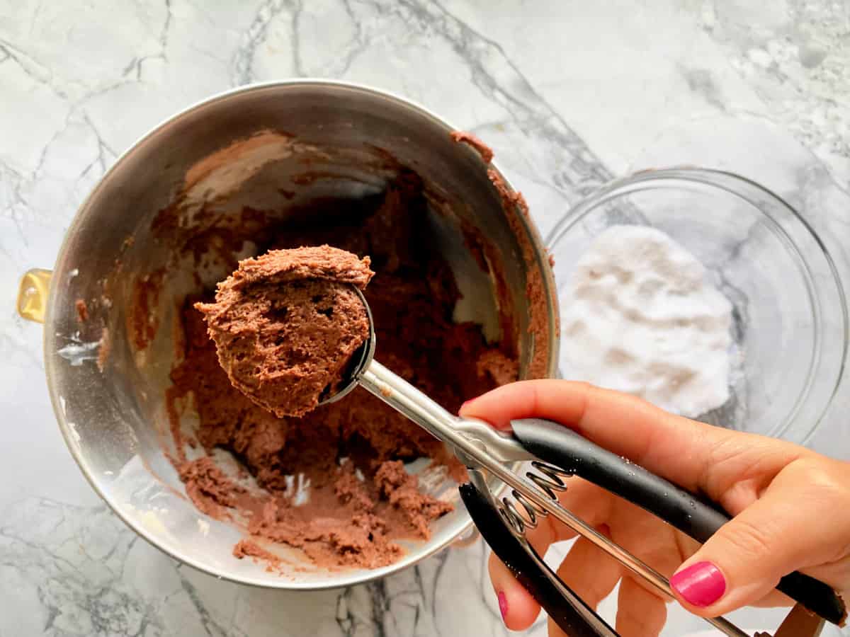 scoop of cookie dough in scoop with mixing bowl with chocolate sugar cookie dough and white sugar in a clear bowl a gray marble counter.