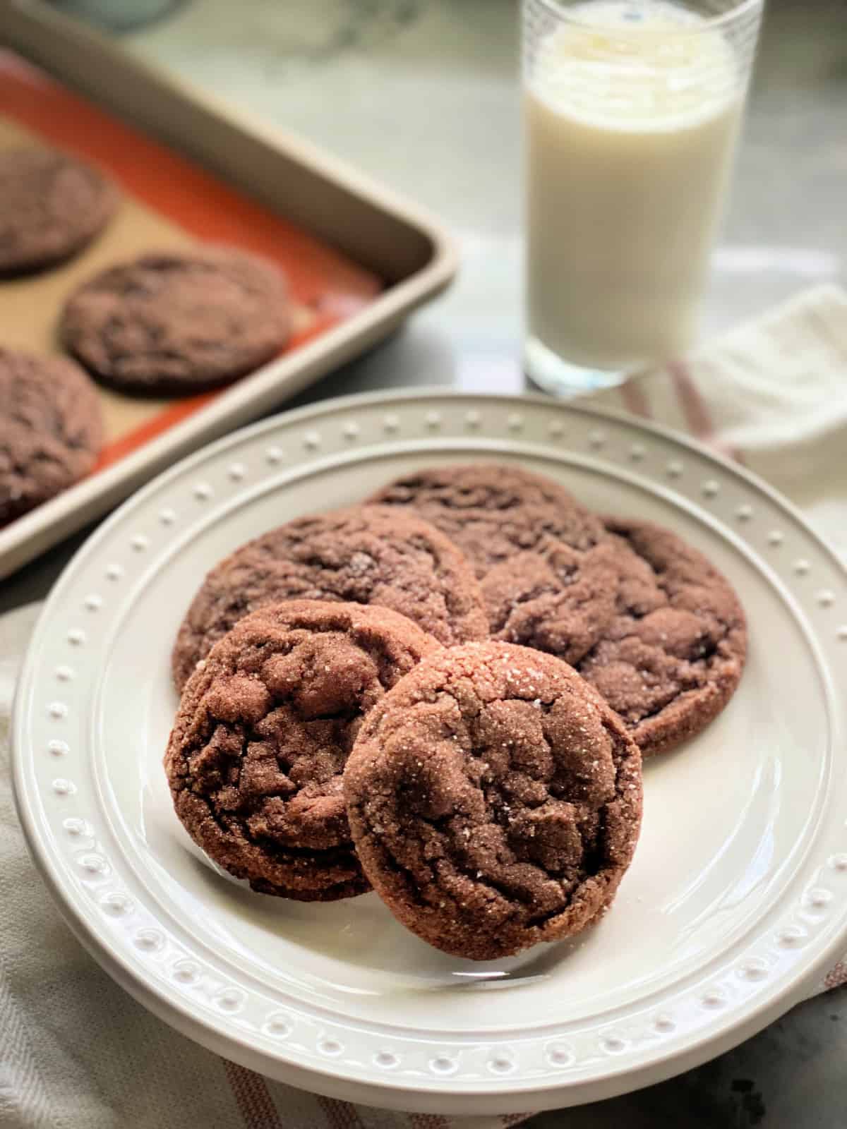 4 chocolate sugar cookies on a white plate with a pink striped white cloth in the background with a glass of milk and 3 baked cookies on a cookie sheet.