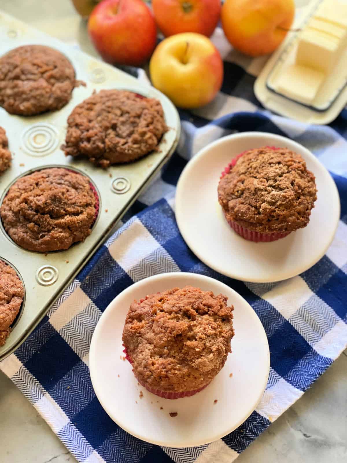 2 apple cinnamon muffins on a small white plates on a blue and white checkered cloth, and 5 muffins, 4 apples and slices of butter in the background.