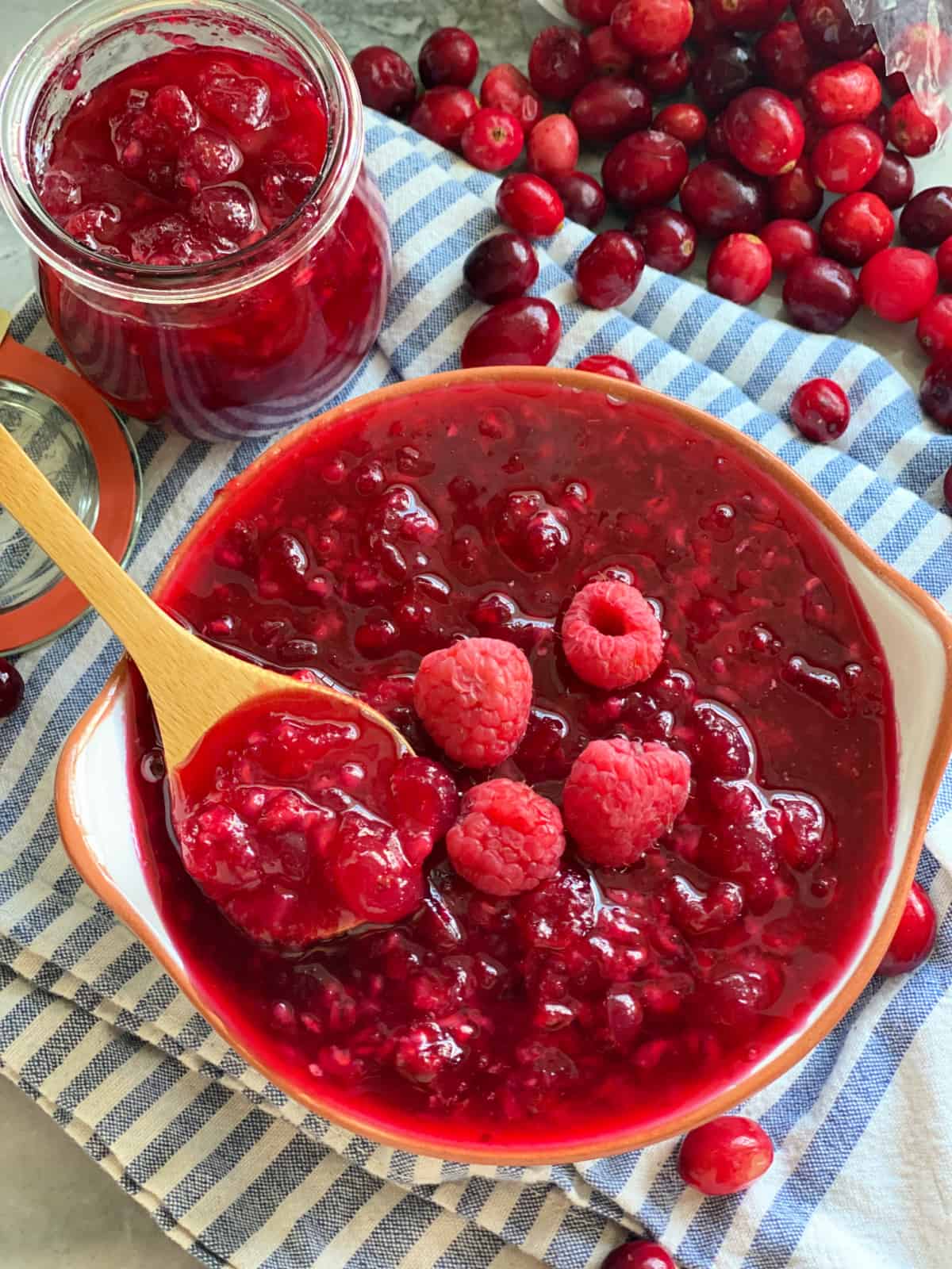 A wooden spoon with cranberry raspberry sauce in a bowl on a blue and white stripped cloth with a jar of sauce and cranberries scattered in the background.