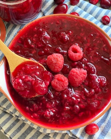 A wooden spoon with cranberry raspberry sauce in a bowl on a blue and white stripped cloth with a jar of sauce and cranberries scattered in the background.