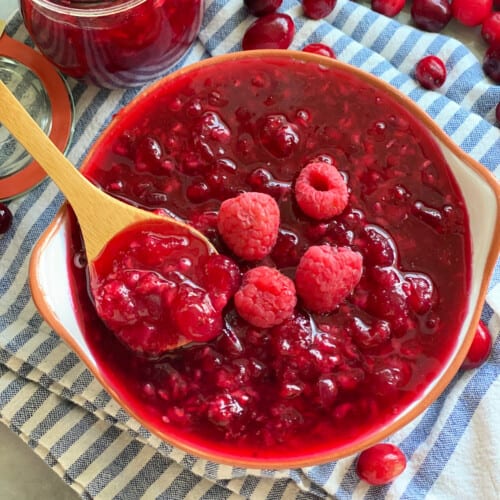 A wooden spoon with cranberry raspberry sauce in a bowl on a blue and white stripped cloth with a jar of sauce and cranberries scattered in the background.