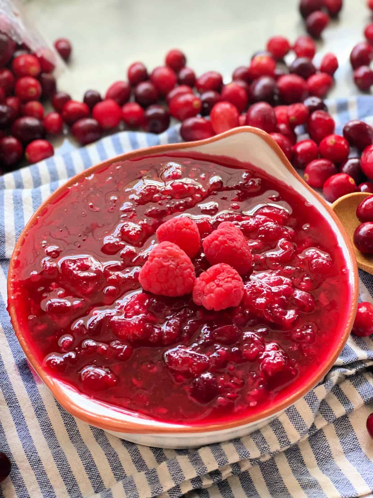 Cranberry raspberry sauce in a bowl on a blue and white stripped cloth with cranberries scattered in the background.