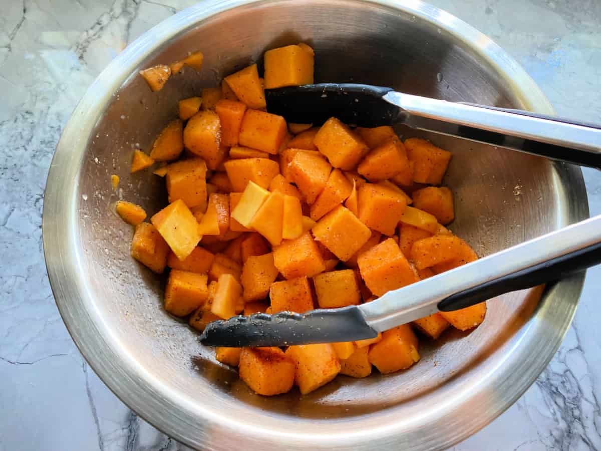 raw diced butternut squash and tongs in a stainless steel bowl on a marble counter.