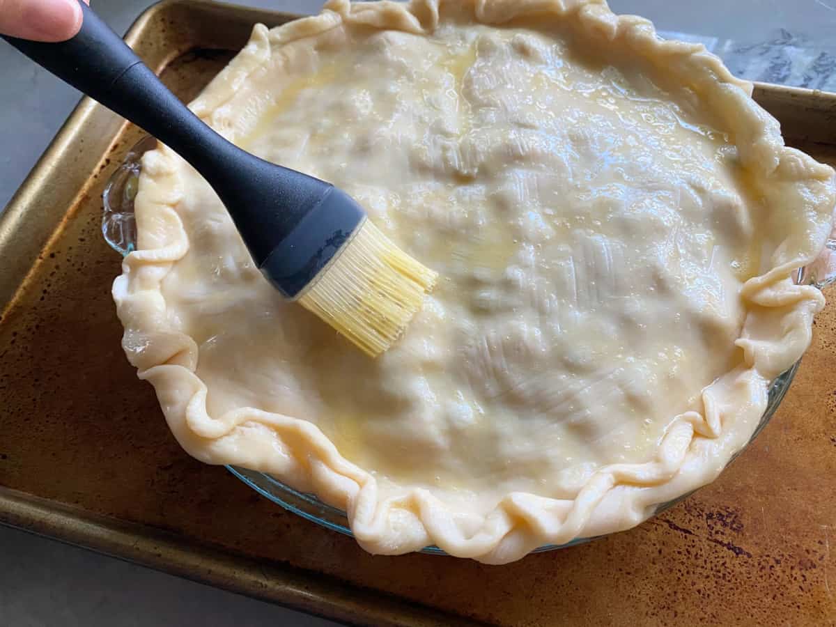 Hand holding a pastry brush, brushing egg wash on a raw pie crust.