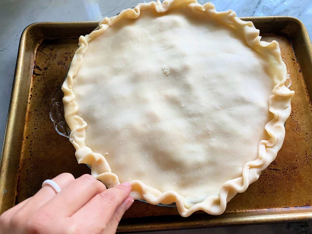 Hand crimping pie crust around a glass pie plate resting on a brown baking sheet.
