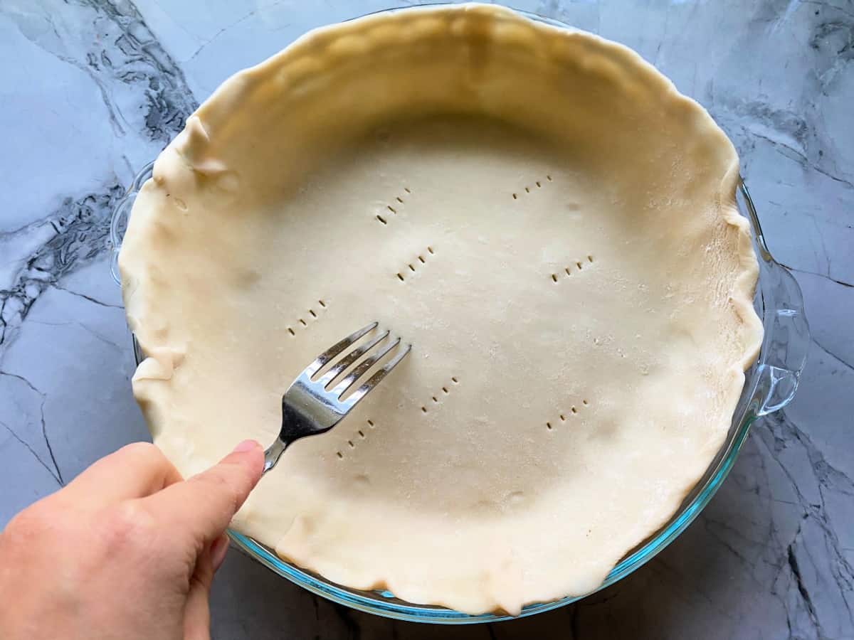 Hand holding a fork and poking on pie dough on a glass pie plate.