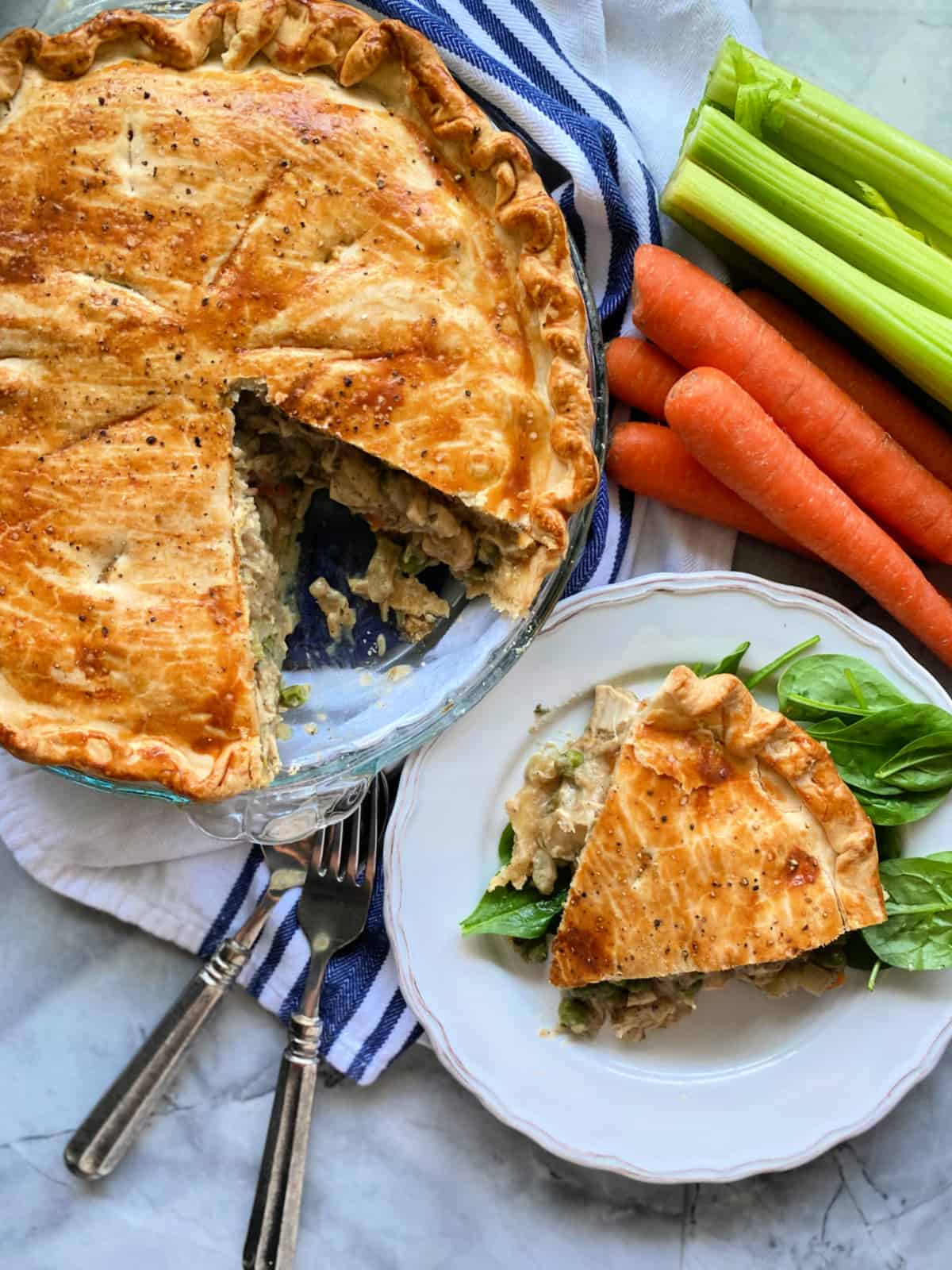 Golden brown baked pie with a slice missing--slice on top of a bed of spinach on a white plate with vegetables next to the pie.