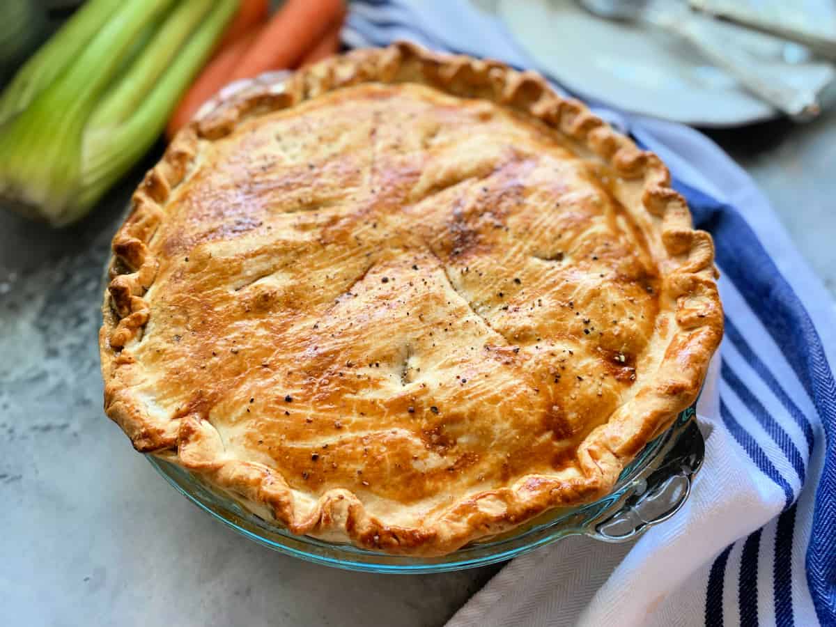 Pie baked in a glass pie dish with white and blue striped cloth next to it and celery and carrots in the background.