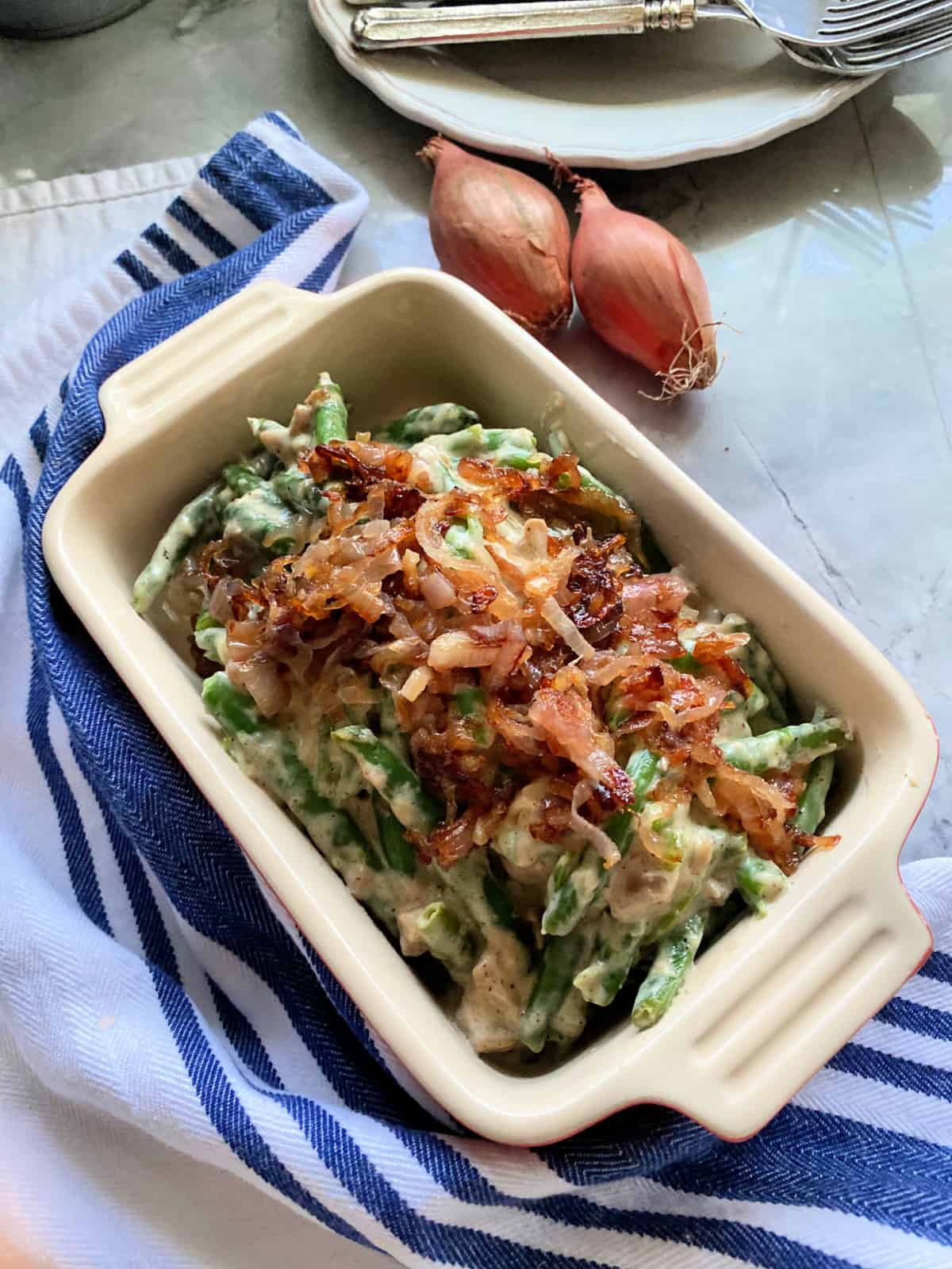 Baking dish with green bean casserole on a blue and white striped cloth with two shallots in the background.