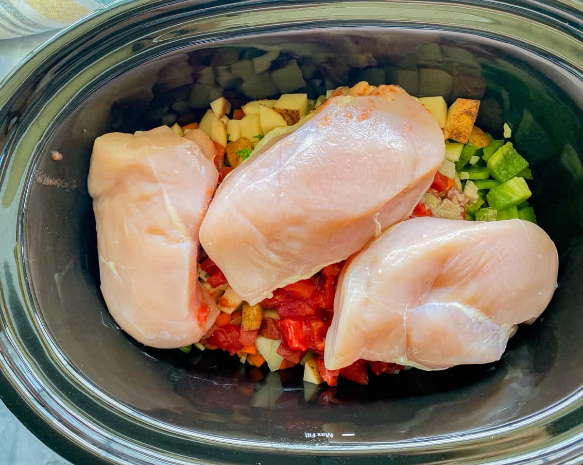 Slow cooker with chopped up ingredients: potato, green pepper, onion, celery, cilantro, carrot, spices, diced tomatoes, and 3 chicken breasts.