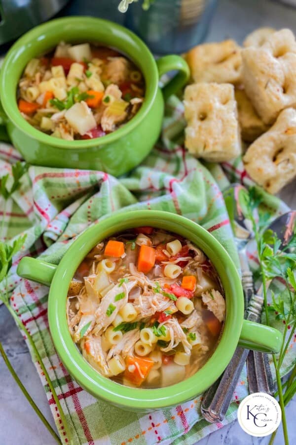 Image with two green bowls with italian chicken soup on a green and pink plaid cloth and cilantro and bread in the background.