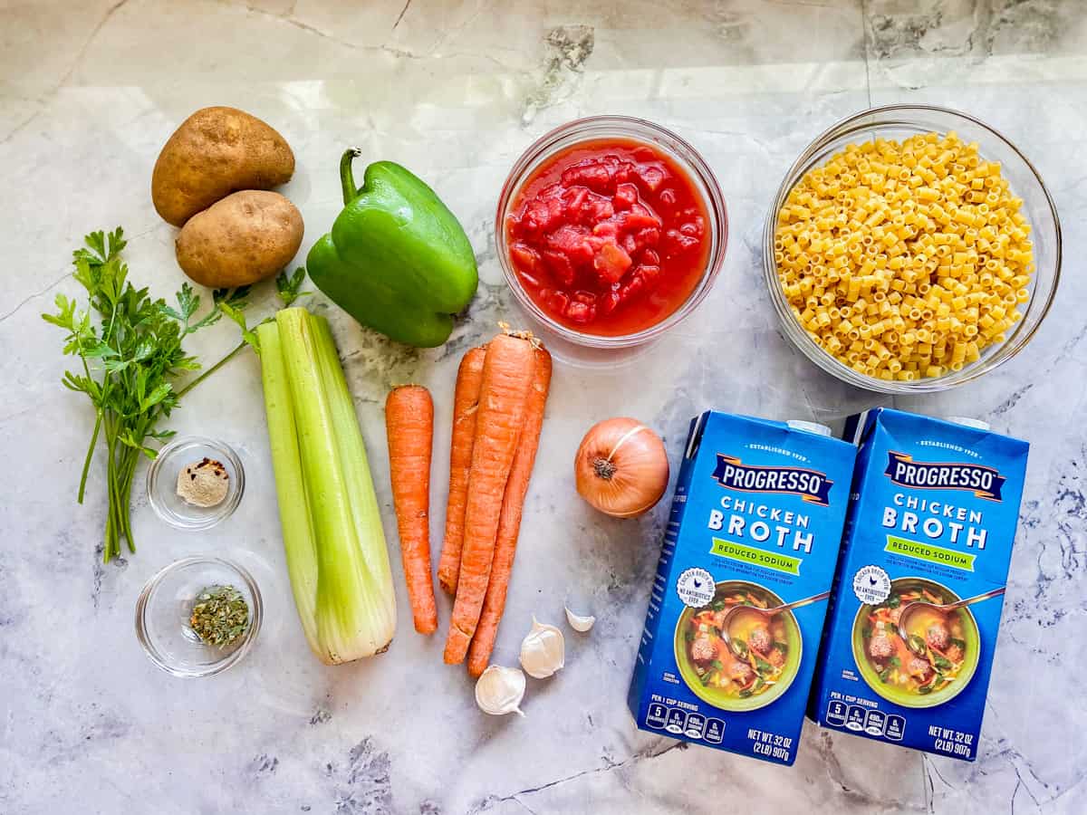 ingredients on marble counter; cilantro, potatoes, spices, celery, carrots, green pepper, garlic cloves, onion, pasta, crushed tomato, and chicken broth