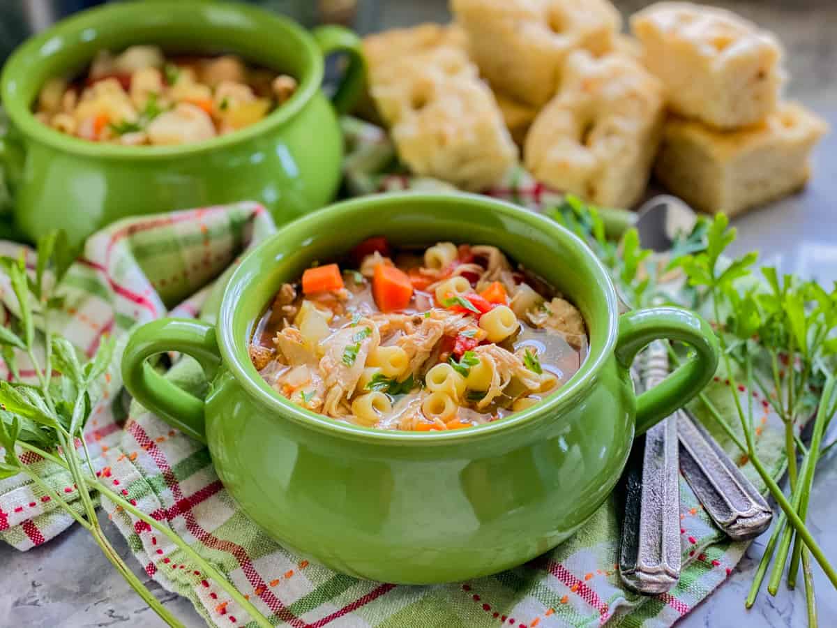 Image with two green bowls with italian chicken soup on a green and pink plaid cloth, two spoons, cilantro, and bread in the background.