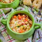 close up of green bowl with italian chicken soup on a green and pink plaid cloth, with two spoons and bread in the background.