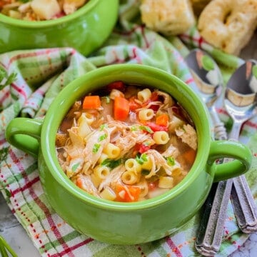close up of green bowl with italian chicken soup on a green and pink plaid cloth, with two spoons and bread in the background.