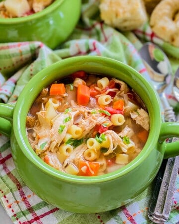close up of green bowl with italian chicken soup on a green and pink plaid cloth, with two spoons and bread in the background.