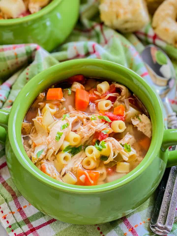 close up of green bowl with italian chicken soup on a green and pink plaid cloth, with two spoons and bread in the background.