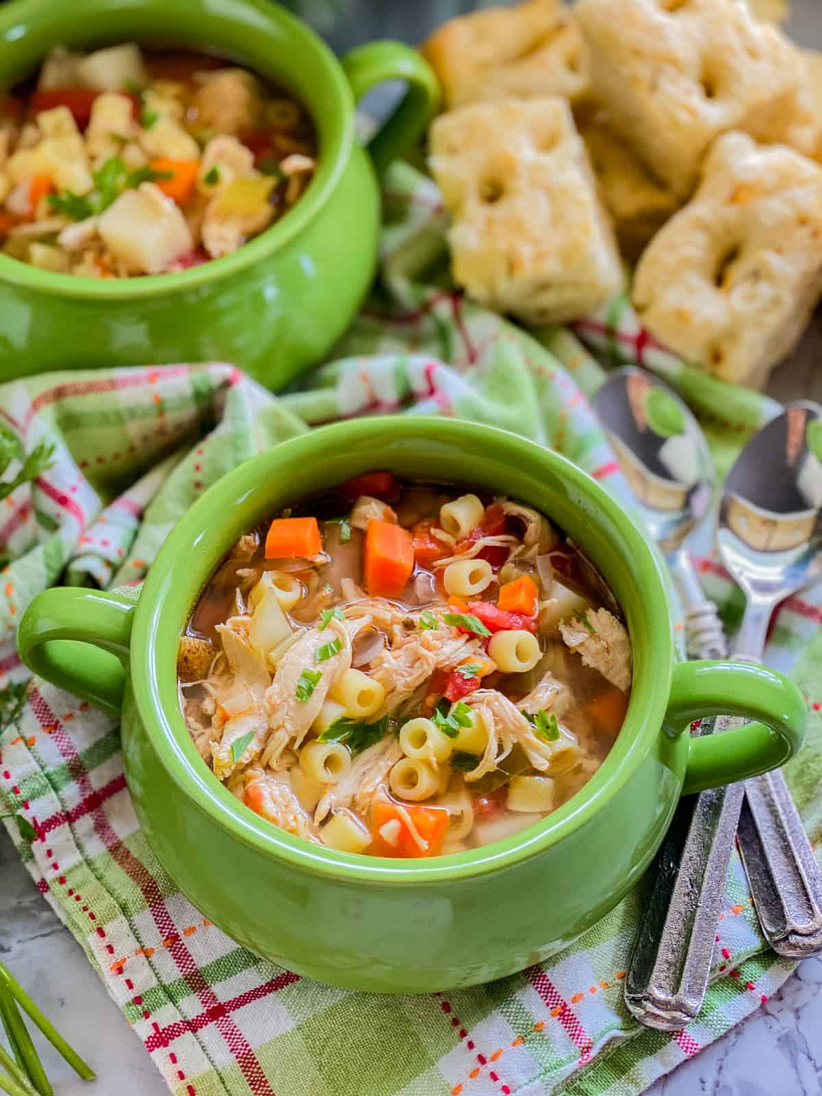 Image with two green bowls with italian chicken soup on a green and pink plaid cloth, two overlapping spoons, and bread in the background.