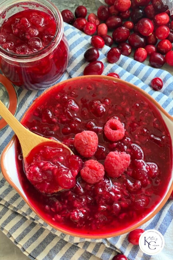 A wooden spoon with cranberry raspberry sauce in a bowl on a blue and white stripped cloth with a jar of sauce and cranberries scattered in the background.