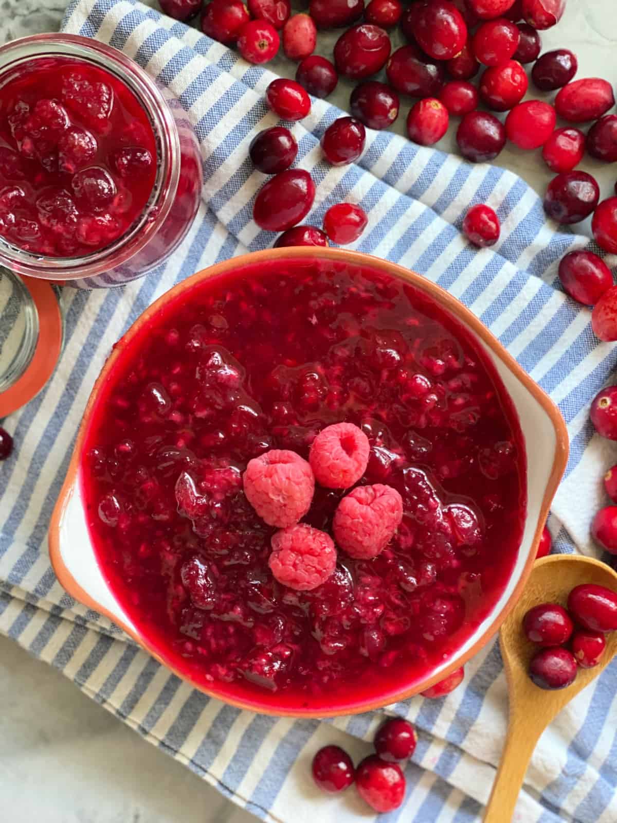 Cranberry raspberry sauce in a bowl on a blue and white stripped cloth with a jar of sauce, wooden spoon, and cranberries scattered in the background.