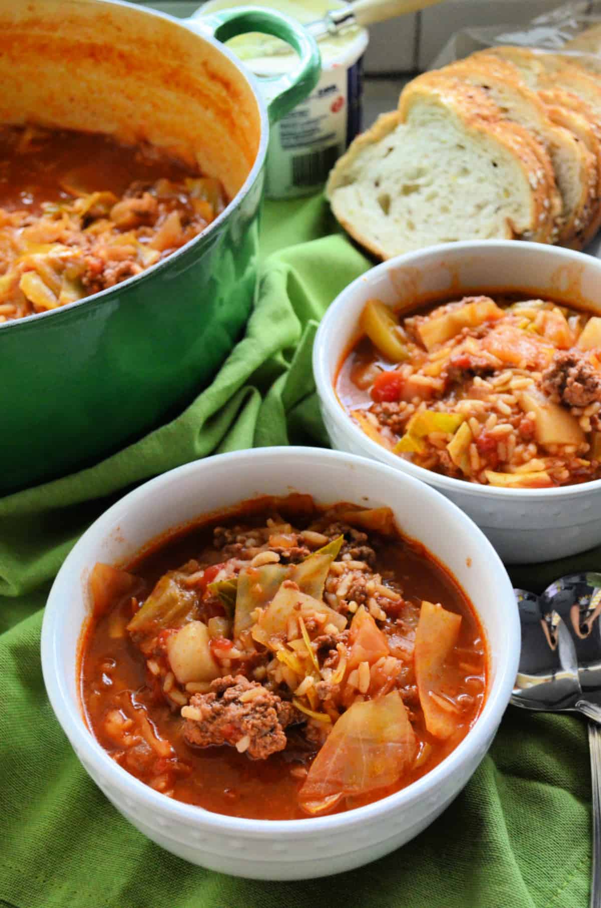 two white bowls with stuffed cabbage soup on green fabric with a pot of soup and slices of bread in the background.