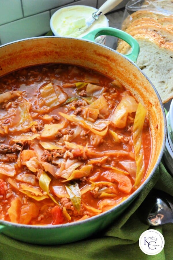 a pot full of cabbage soup on green fabric and slices of bread and white bowls in the background.